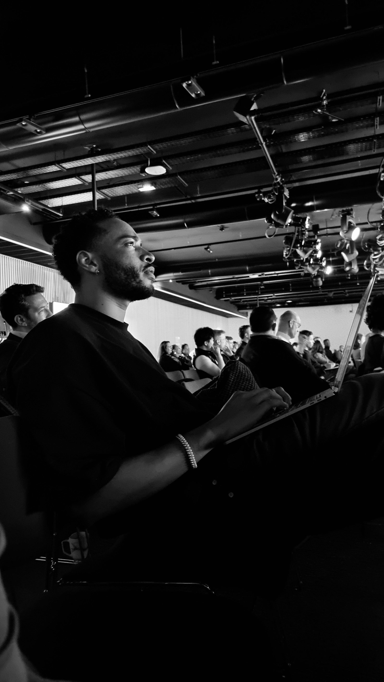 Swan Badol assis dans une salle, un ordinateur portable sur les genoux, le public et les projecteurs derrière lui.